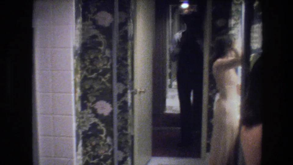 8mm home-movie frame circa 1977: woman in cream gown and man in dark clothing holding a camera stand in a wallpapered interior hallway beside a large mirror with overhead light visible.