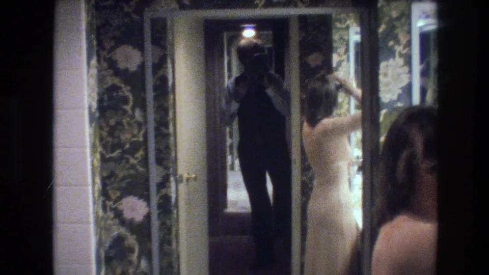 8mm frame circa 1977: mirror-within-shot composition with the cameraperson's dark silhouette holding a camera reflected in the mirror, woman in cream gown in foreground, dark floral wallpaper behind.