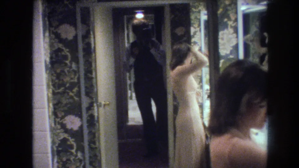 Final 8mm frame circa 1977: woman in cream gown in partial profile before a mirror, cameraman's dark silhouette with camera reflected behind her, dark floral wallpaper on either side.