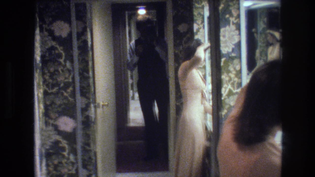 Woman in cream chemise stands before a large mirror in a dimly lit bathroom with floral wallpaper; camera operator's silhouette is reflected behind her.