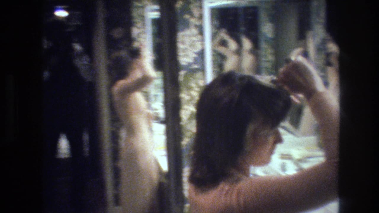 Woman seen from behind styling dark hair with hands raised near a multi-panel mirror showing her reflection; floral wallpaper surrounds the scene.