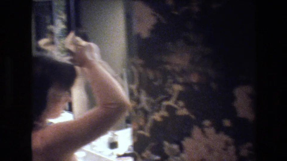 8mm home movie still, circa 1977 — tight handheld shot of woman styling dark hair, light-colored garment at shoulder, bathroom counter with small containers below and floral wallpaper at right.