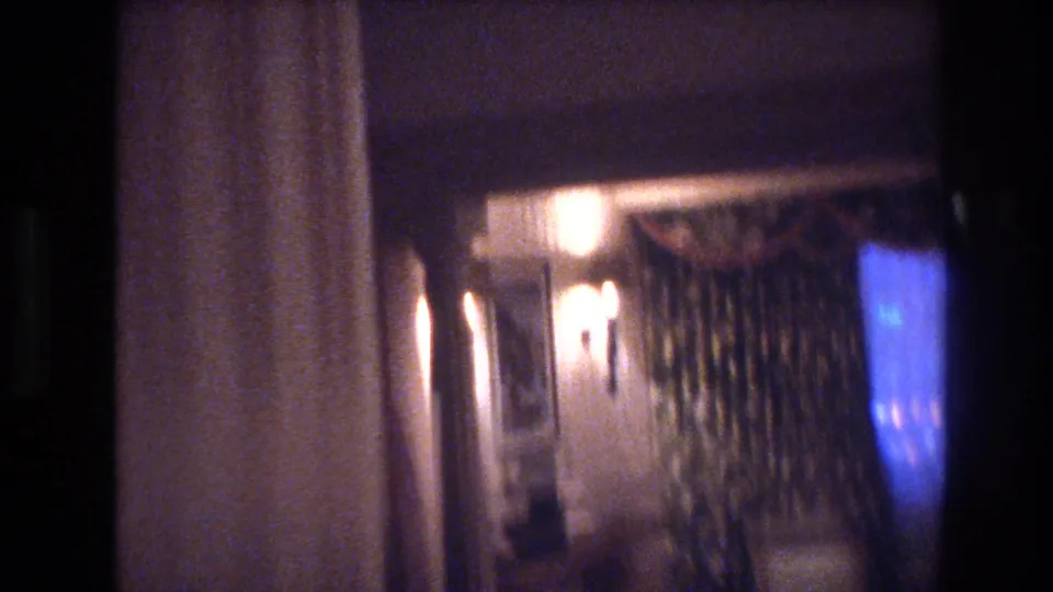 8mm home movie still, circa 1977 — camera pans to a dimly lit room with warm wall-sconce lighting, heavy drapes, and a bluish glow at right suggesting a television screen.