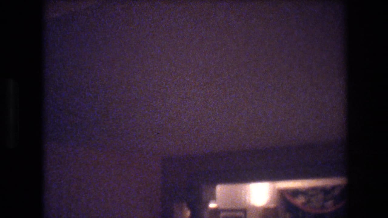 Heavily underexposed 8mm hotel room interior, Las Vegas, circa 1977, showing a warm lamp and dark curtain before the camera turns toward the window.