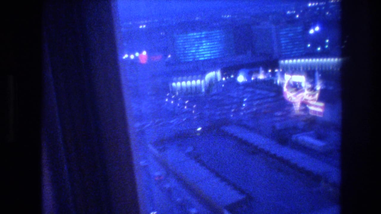 Blue-shifted 8mm nighttime view of the Las Vegas Strip from a hotel window, circa 1977, with neon-lit casino buildings and a decorative lit structure visible in the midground.