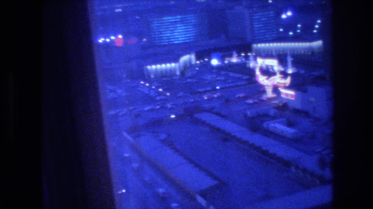 8mm nighttime panorama of the Las Vegas Strip from a hotel window, circa 1977, with blue color shift, heavy grain, and a large illuminated structure visible at street level.