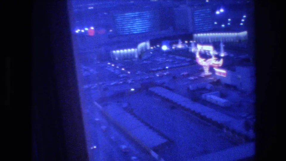 8mm nighttime panorama of the Las Vegas Strip from a hotel window, circa 1977, with blue color shift, heavy grain, and a large illuminated structure visible at street level.