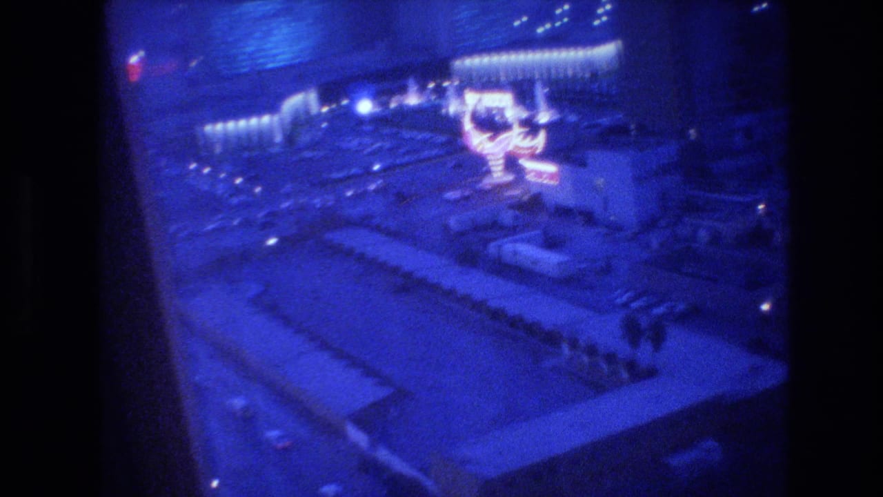 Closing 8mm frame of the Las Vegas Strip at night from a hotel window, circa 1977, blue-shifted and grain-heavy, with lit casino structures and street below.