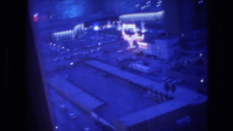 Closing 8mm frame of the Las Vegas Strip at night from a hotel window, circa 1977, blue-shifted and grain-heavy, with lit casino structures and street below.
