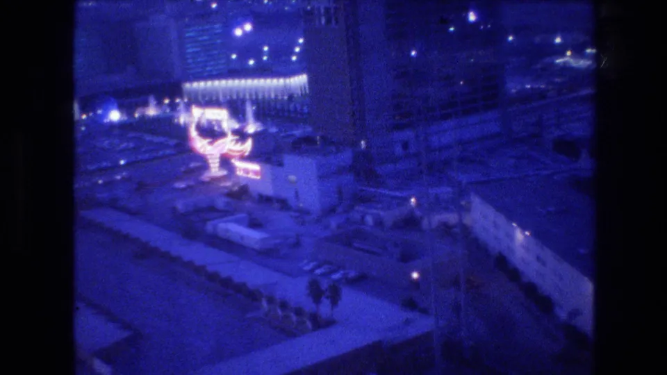 Shifted handheld view from upper floor over Las Vegas at night, circa 1977, revealing surrounding cityscape, a neon sign, and tiny figures at street level.