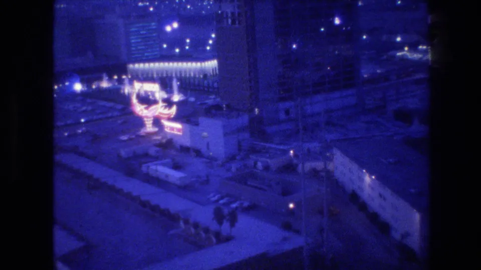 Closing frame of a handheld 8mm panning shot over Las Vegas at night, circa 1977, with a neon sign and surrounding city lights extending to the horizon.
