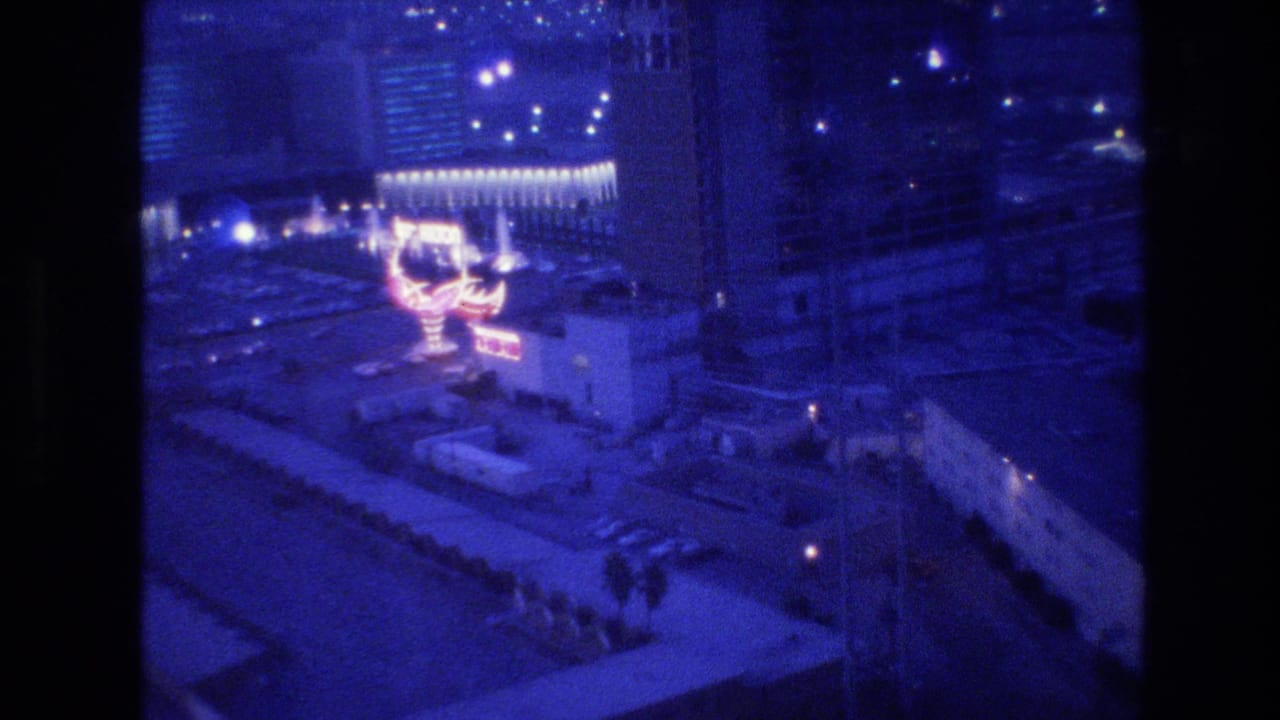 8mm film frame circa 1977, aerial view of Las Vegas at night from a hotel high-rise, showing a large pink-red neon sign and pedestrian-scale figures on the street below.