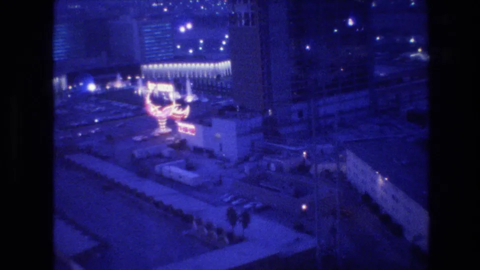 8mm film frame circa 1977, aerial view of Las Vegas at night from a hotel high-rise, showing a large pink-red neon sign and pedestrian-scale figures on the street below.