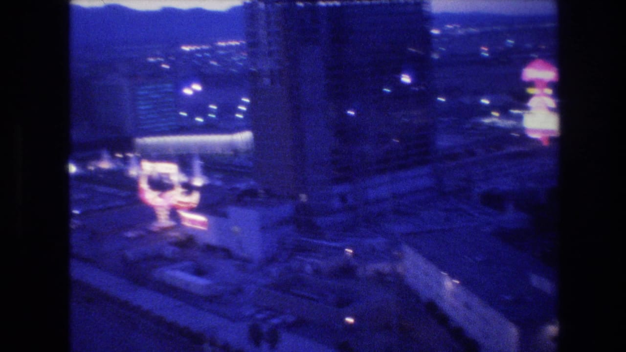 8mm home movie frame circa 1977, Las Vegas Strip at night showing a high-rise hotel at left and a prominent domed pink neon structure at right, with mountain ridgeline on the horizon.