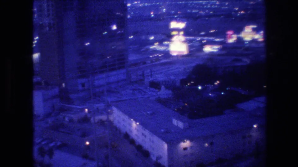 8mm film frame circa 1977, wide aerial view of Las Vegas at night from hotel high-rise, neon sign cluster glowing pink in middle distance, low-rise rooftops in foreground.