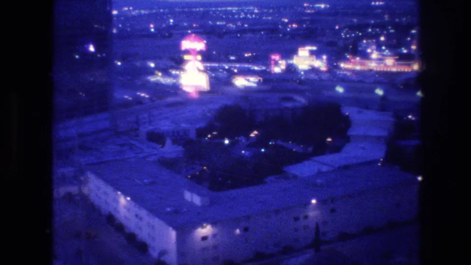 8mm home movie frame circa 1977, Las Vegas Strip at night with a distinctive rounded pink neon sign structure prominent at center and a tall hotel building at right.
