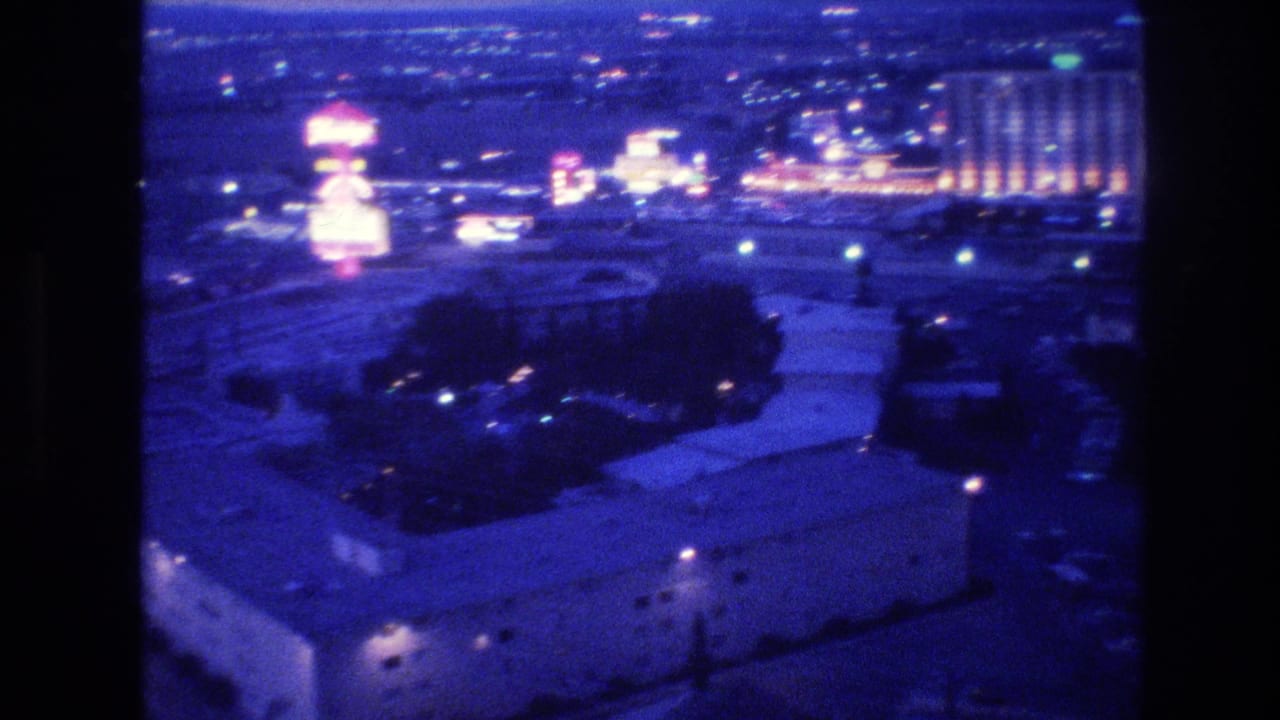 Elevated nighttime view of Las Vegas showing low-rise buildings and a distant cluster of illuminated hotel towers under a heavy blue-purple 8mm color cast.