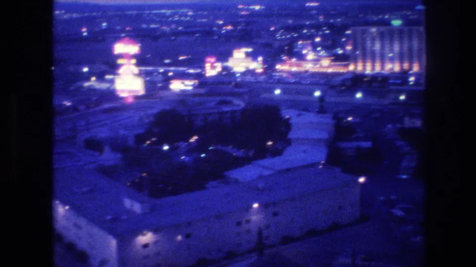 8mm frame from 1977 Las Vegas: elevated night pan with a neon-lit hotel cluster glowing on the horizon and flat-roof buildings filling the foreground.
