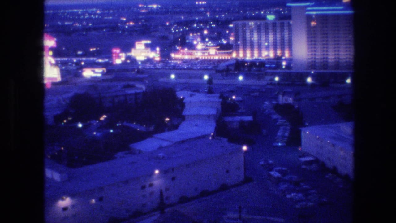 Mid-pan 8mm frame of Las Vegas at night, circa 1977, with large lit hotel towers emerging on the right side against a dark sky and blue-tinted rooftops below.