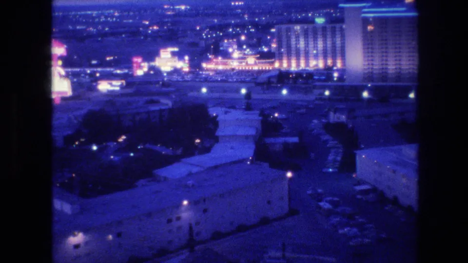 Mid-pan 8mm frame of Las Vegas at night, circa 1977, with large lit hotel towers emerging on the right side against a dark sky and blue-tinted rooftops below.