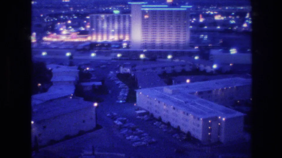 8mm frame circa 1977 showing a tall Las Vegas hotel tower with a lit-window grid and cyan roofline accent light, parking areas and low-rise buildings in the blue-purple-toned foreground.