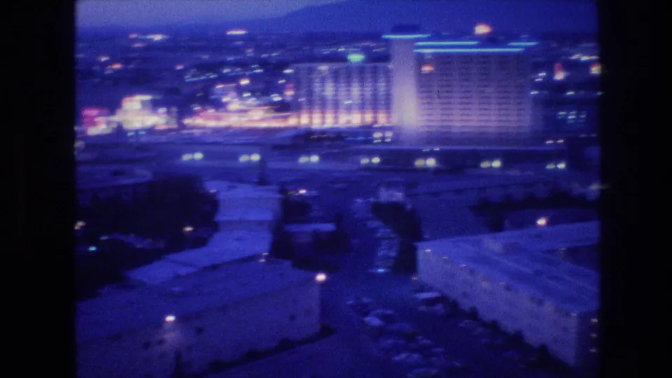 Final 8mm pan frame of Las Vegas at night circa 1977, hotel tower right of center over an expanse of surface parking and low-rise structures rendered in blue-purple tones.
