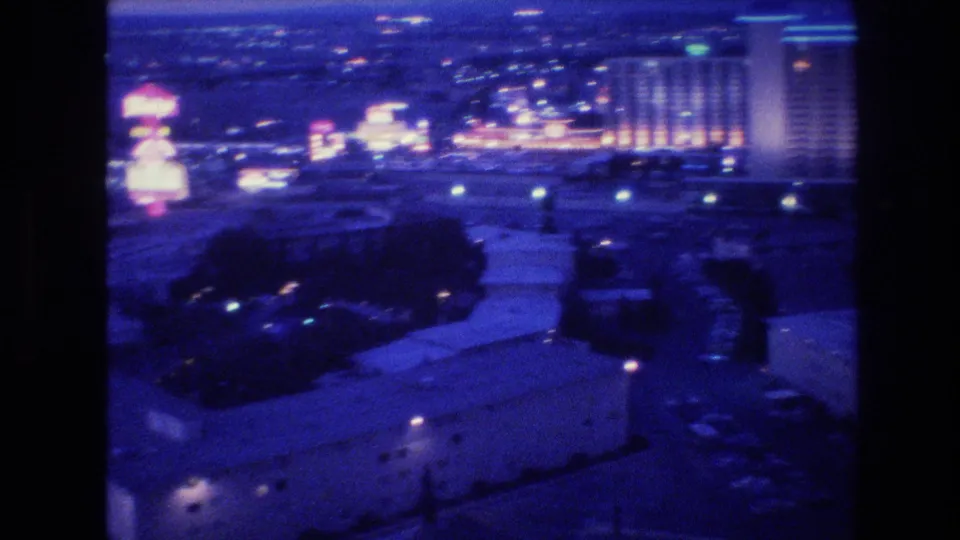 1977 Las Vegas nighttime exterior view from a high vantage point, with glowing hotel-casino buildings, neon lights, dark roads, and soft 8mm color.