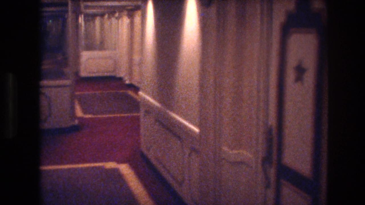 1977 Las Vegas hotel hallway with red carpeting, cream-colored paneled walls, warm ceiling lights, and a softly exposed home-movie view down the corridor.