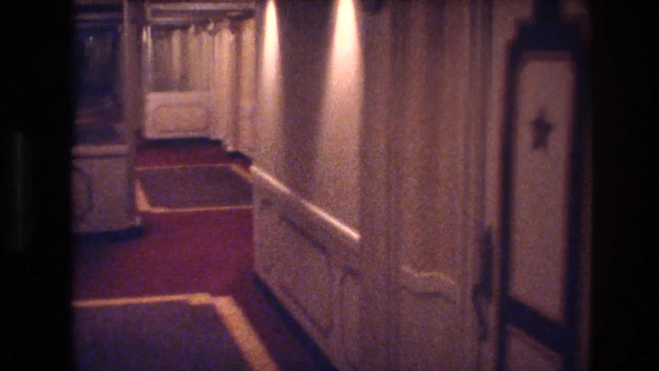 1977 Las Vegas hotel hallway with red carpeting, cream-colored paneled walls, warm ceiling lights, and a softly exposed home-movie view down the corridor.