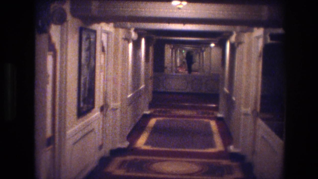 Long hotel corridor with patterned carpet and paneled walls; small figure visible in the distance.