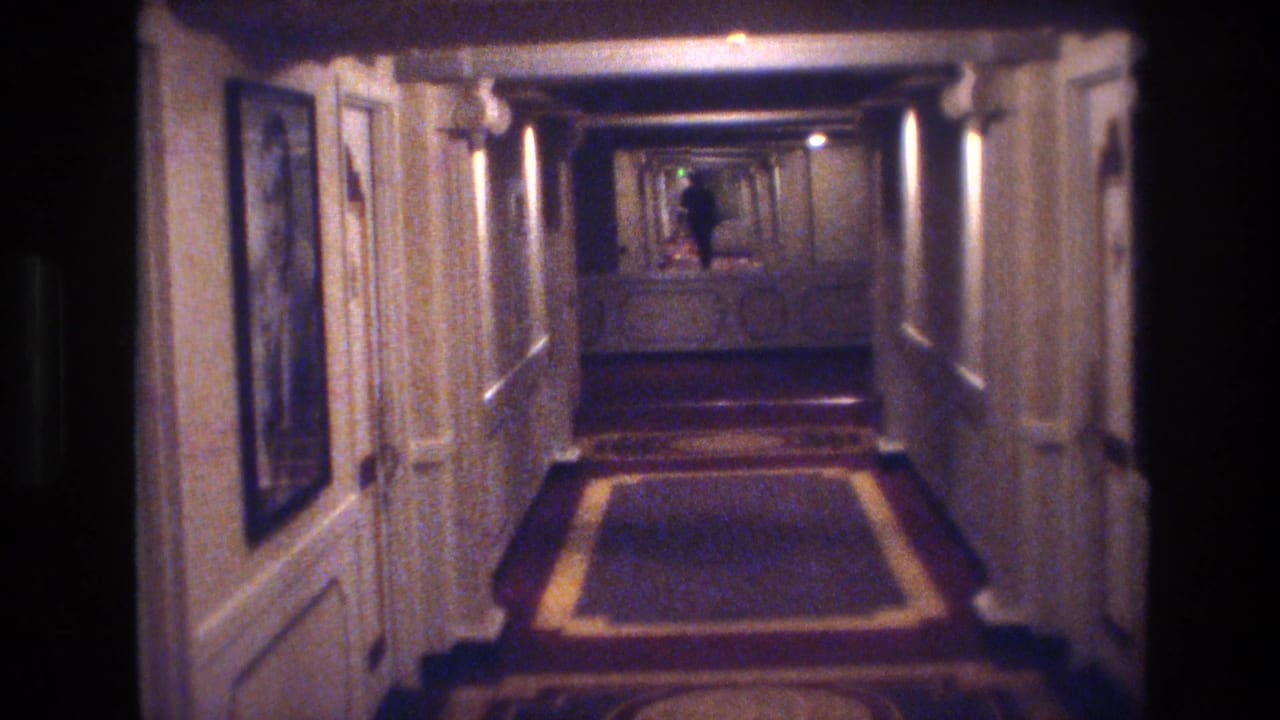 Hotel corridor with ornate carpet; figure visible at the far end under ceiling lights.
