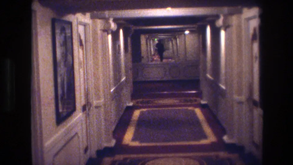 1970s Las Vegas hotel hallway filmed on 8mm; paneled walls, geometric carpet runner, and a figure at the corridor end under warm overhead lighting.