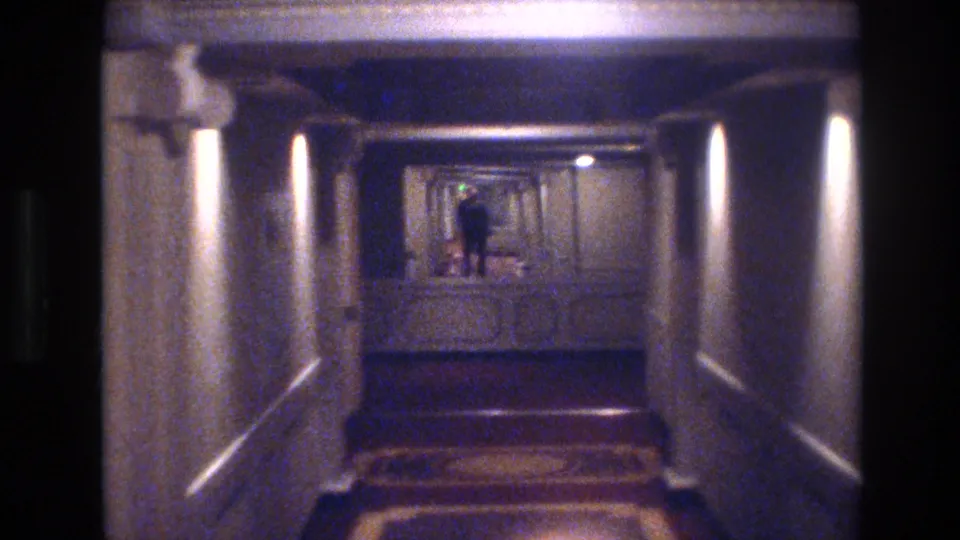 1970s Las Vegas hotel corridor on 8mm; wall-mounted sconces cast pools of light along paneled walls as a figure stands in the middle distance.