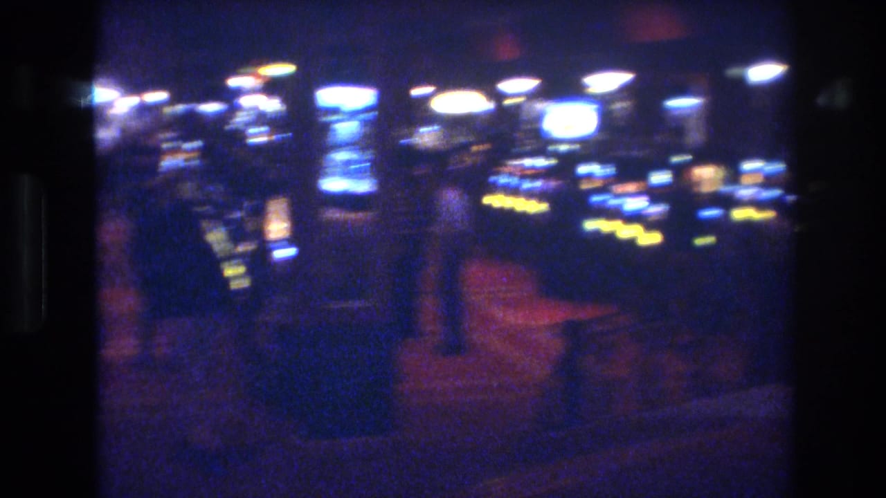 Dimly lit casino interior with rows of illuminated slot machines and a blurred figure walking the aisle.