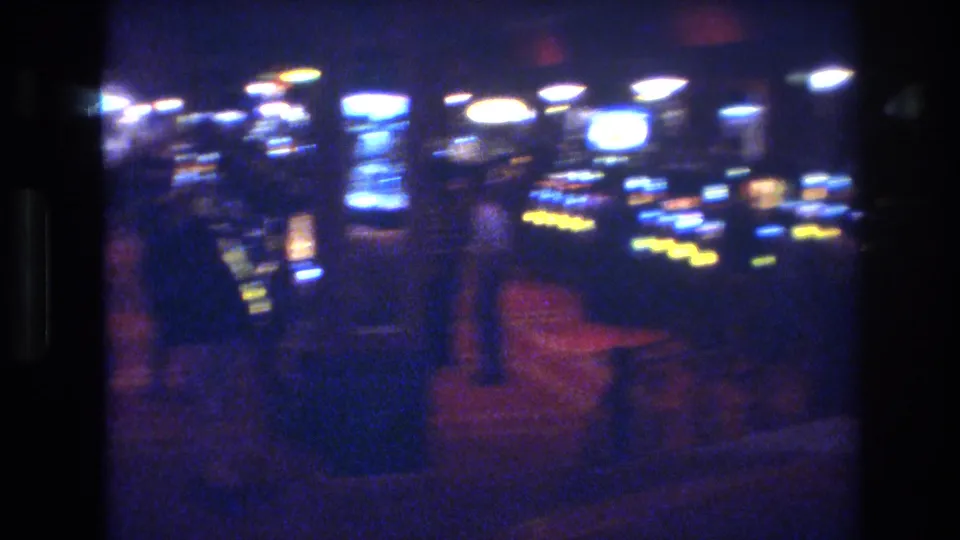 8mm film frame from a 1977 Las Vegas casino showing slot machines with yellow and blue illuminated panels and a blurred patron figure in the foreground.