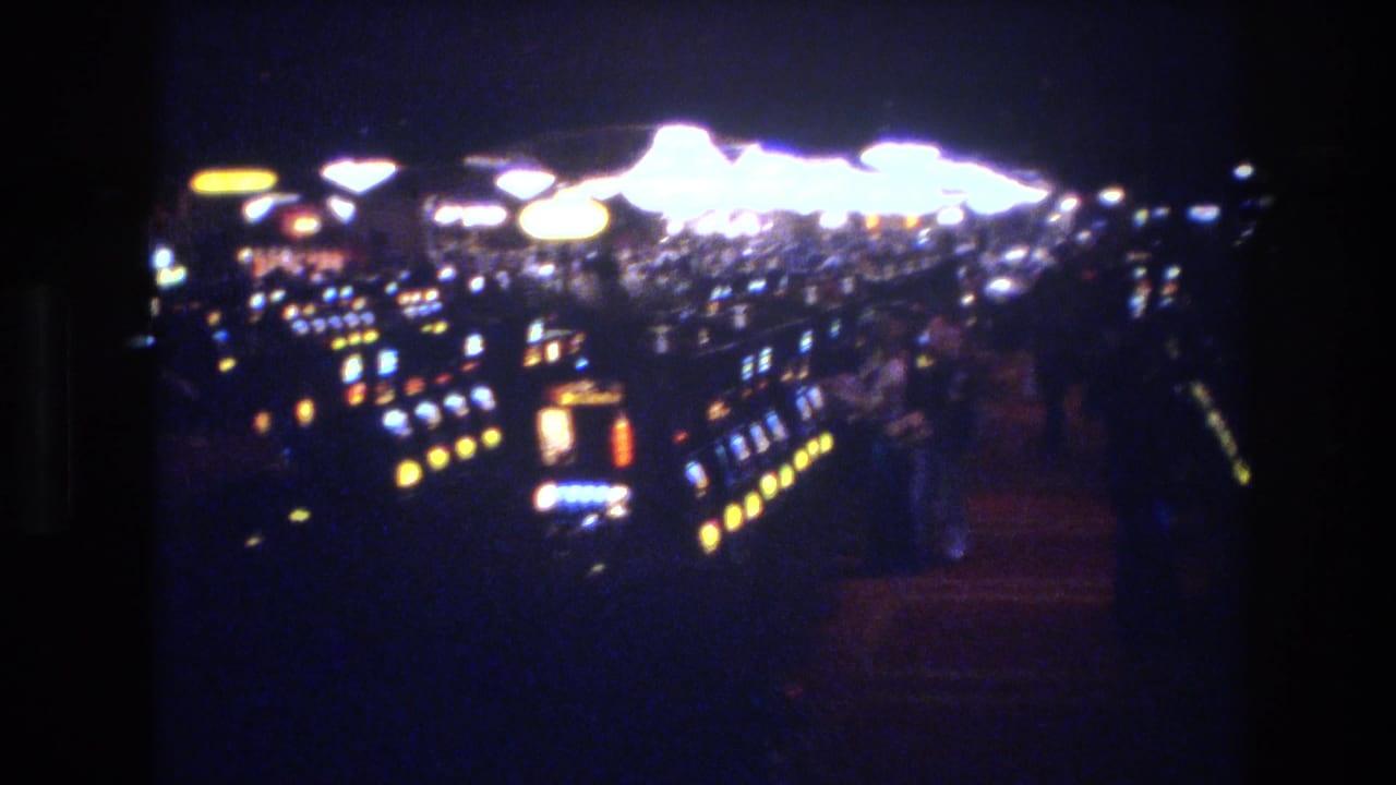 8mm film view across long rows of slot machines in what appears to be a 1970s Las Vegas casino, with large round overhead light fixtures visible above.