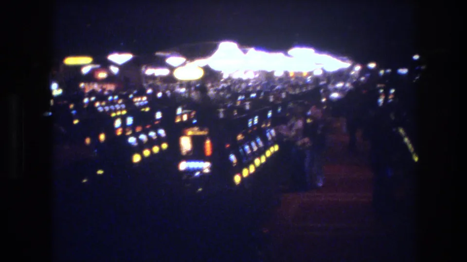 8mm film view across long rows of slot machines in what appears to be a 1970s Las Vegas casino, with large round overhead light fixtures visible above.