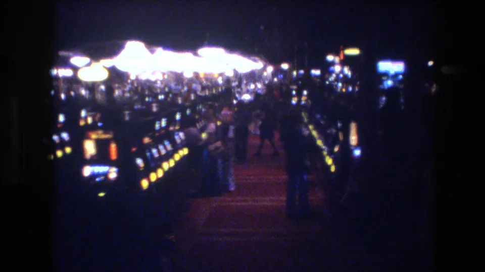 8mm film frame showing a busy 1970s Las Vegas casino floor with multiple blurred patrons among long rows of slot machines and large overhead light fixtures.
