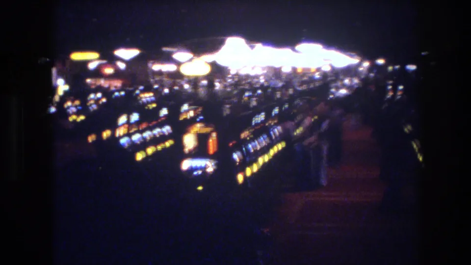8mm film frame showing slot machine rows receding into darkness under large round overhead lights in what appears to be a 1970s Las Vegas casino.