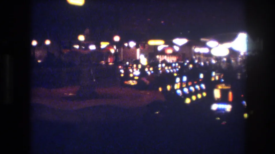 8mm footage of a Las Vegas casino floor, circa 1977, with slot machine banks extending into shadow and yellow coin-tray lights arrayed in rows against a dark background.