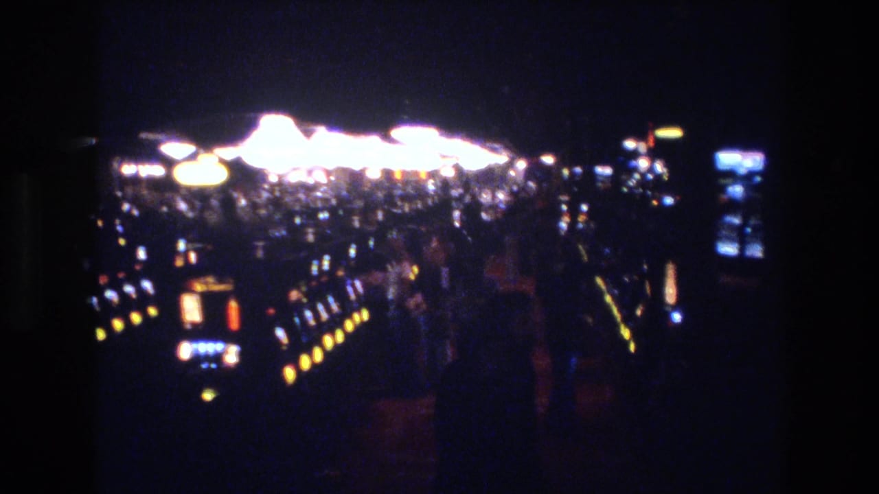 8mm home-movie frame from a Las Vegas casino, circa 1977, showing a panoramic sweep of slot machine banks with yellow coin-tray lights and a dense cluster of blurred overhead illumination.