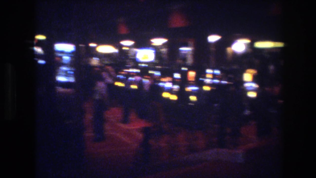 8mm footage from a Las Vegas casino, circa 1977, showing slot machine cabinets with yellow coin lights and blue displays, and indistinct figures of patrons visible at right against a red-toned carpet.
