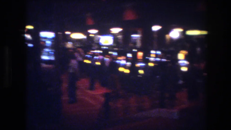 8mm footage from a Las Vegas casino, circa 1977, showing slot machine cabinets with yellow coin lights and blue displays, and indistinct figures of patrons visible at right against a red-toned carpet.