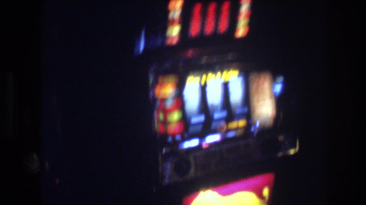 Illuminated 1970s slot machine in a dark casino, showing spinning white reels and colored lights above, with a yellow lion logo visible below.
