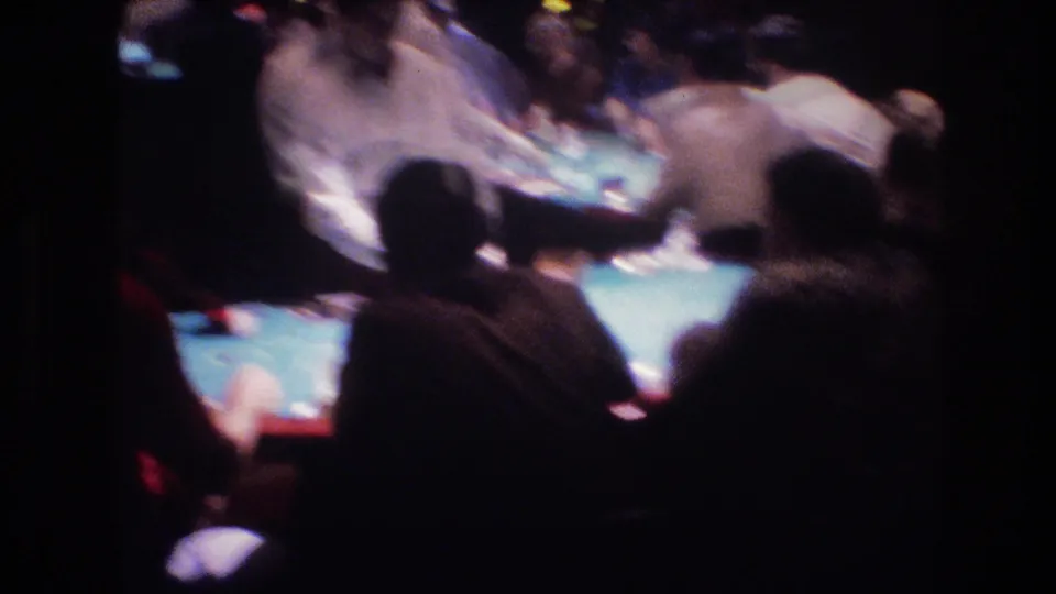8mm footage of patrons crowded around a green felt card table in a low-light Las Vegas casino, circa 1977; figures seen from behind with no legible faces.