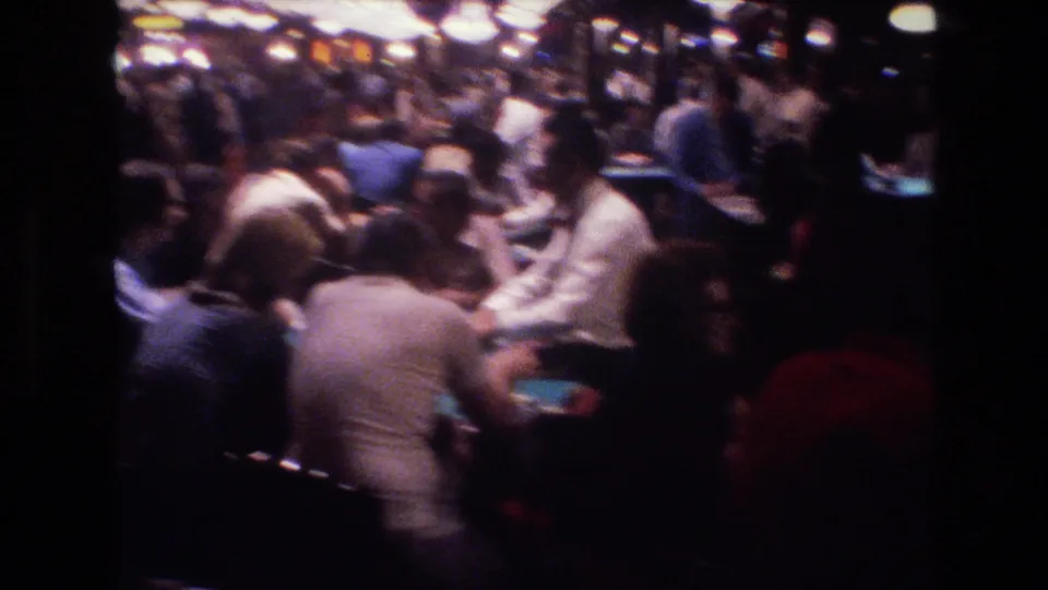 Handheld 8mm shot of a packed Las Vegas casino floor in 1977, patrons clustered at card tables under dim overhead globe lights.