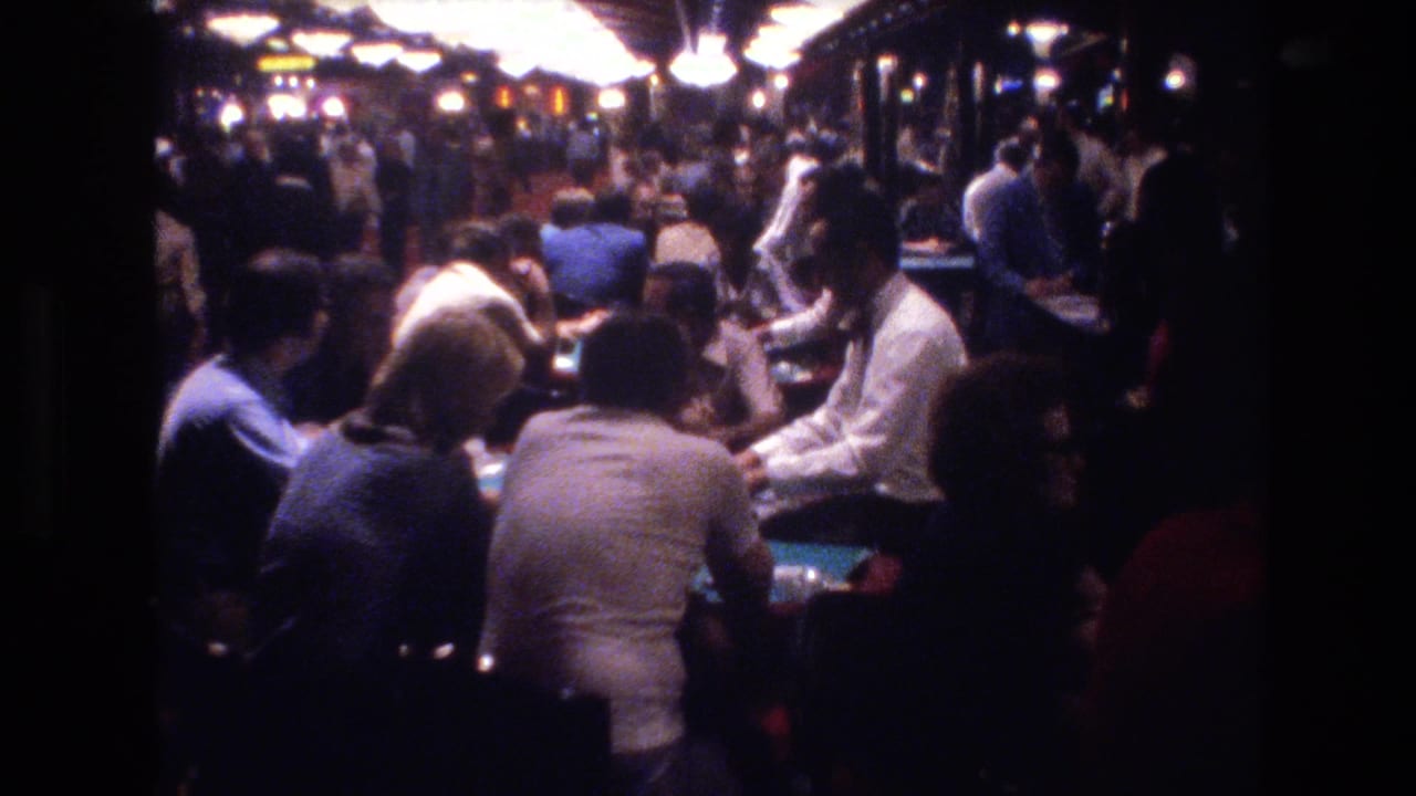 Casino floor crowd at a slightly different angle, players around tables under overhead globe lights with colorful background signage.