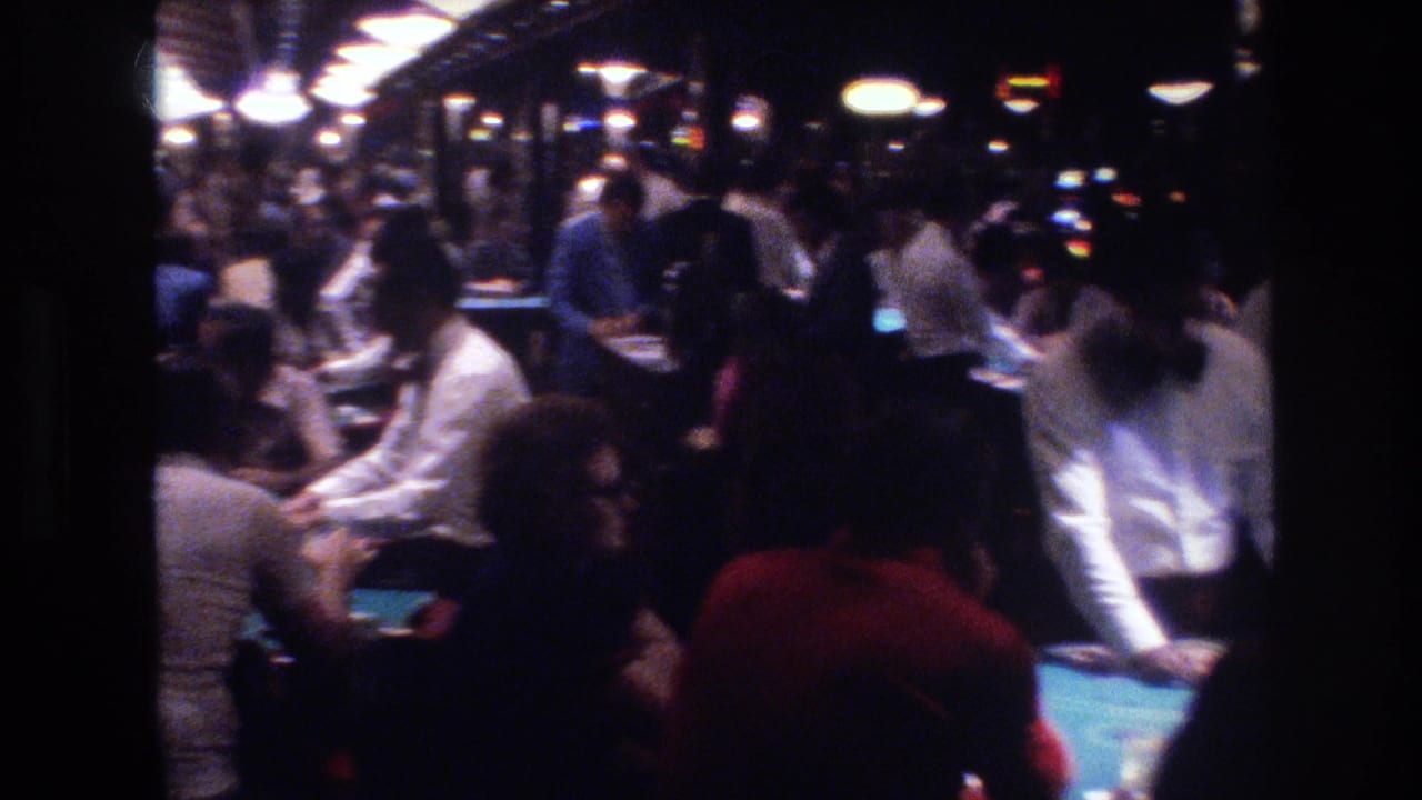 8mm wide shot of Las Vegas casino interior circa 1977, showing the full depth of the gaming floor, pendant globe lights, and patrons at multiple tables.