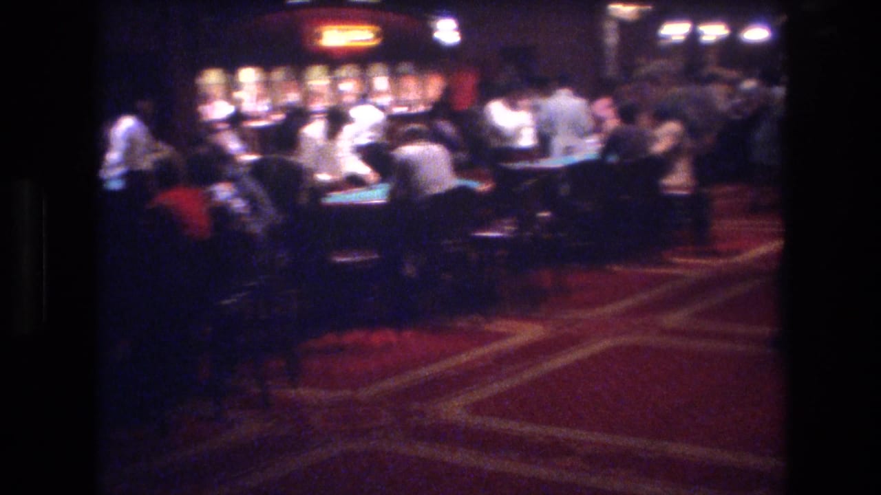 Super 8 wide shot of a Las Vegas casino floor circa 1977, showing rows of gaming tables, red patterned carpet, and patrons in casual dress under incandescent lighting.