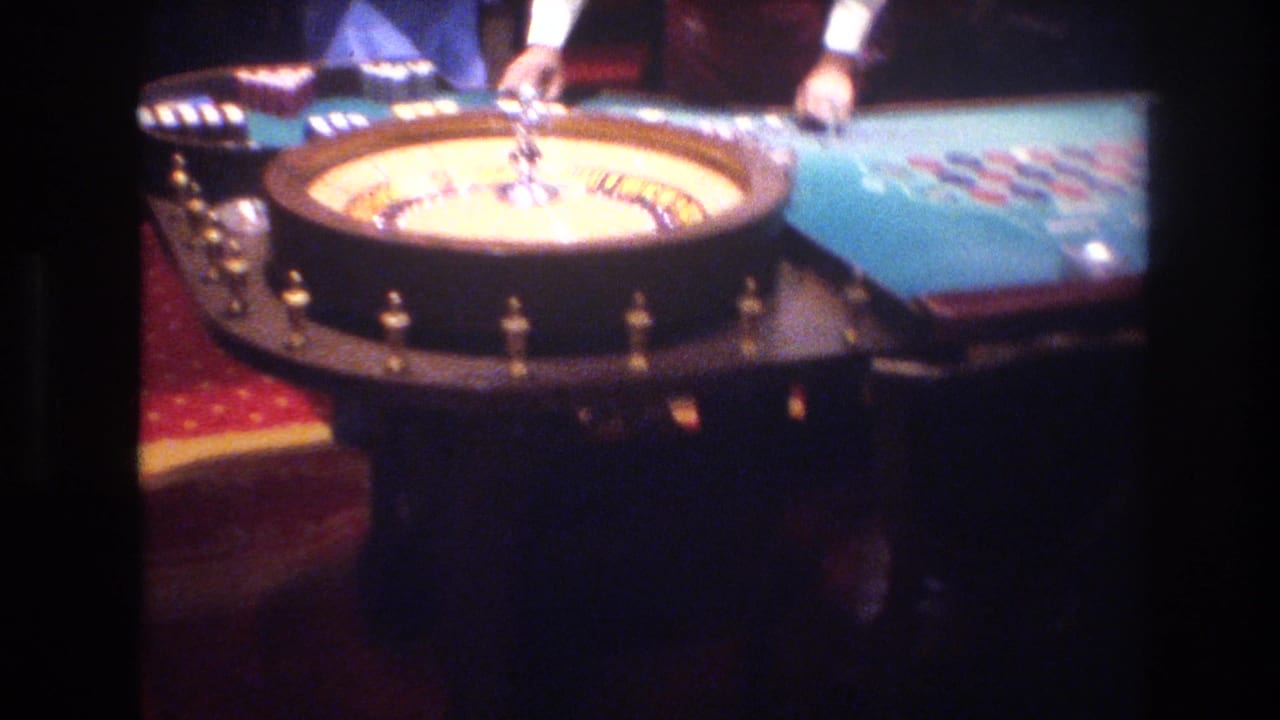 Super 8 close-up of a roulette wheel at a Las Vegas casino table circa 1977, with blue felt betting layout, stacked chips, and a dealer's forearms at frame edge.